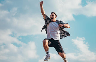 A joyful young man is leaping with a big smile against a background of blue sky and fluffy white clouds.
