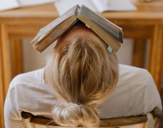 A woman is stressed and she is sitting on the chair with a book on her head.