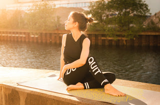 A woman sitting outside during sunset, practicing yoga and self care. She wears black sport outfit, and bare feet. She is sitting next to the river. This image is related to self care during easer holiday season.