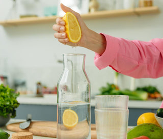 A hand in pink shirt squeezing a lemon into a glass clear jar with water. The jar is placed in the kitchen counter among other fruits, herbs and vegetables.