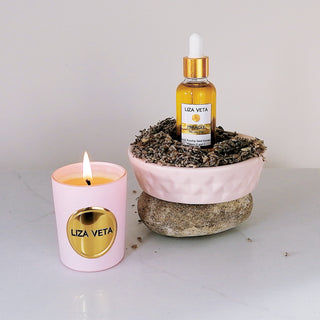 A small pink candle, made of natural wax and scented with essential oil, sits next to a glass bottle of natural face oil on a grey stone. Dry lavender is placed beside the items.