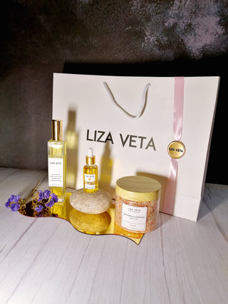 This is a gift set that includes room and linen spray in a glass square bottle, natural face oil and bath salts in a transparent jar. There is a purple flower next to it. This image is related to how to choose a perfect gift set.
