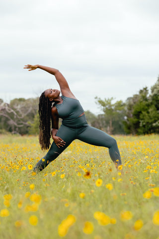A black woman in green grey sport outfit practicing aerobics outside in a field with yellow flowers. This image is related to self care during spring.