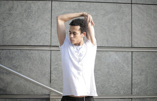 A man wearing a white t-shirt is performing stretches in a work environment with a grey wall as the background. He appears to be listening to music while stretching.